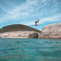 man doing a flip off a rock into the beach water